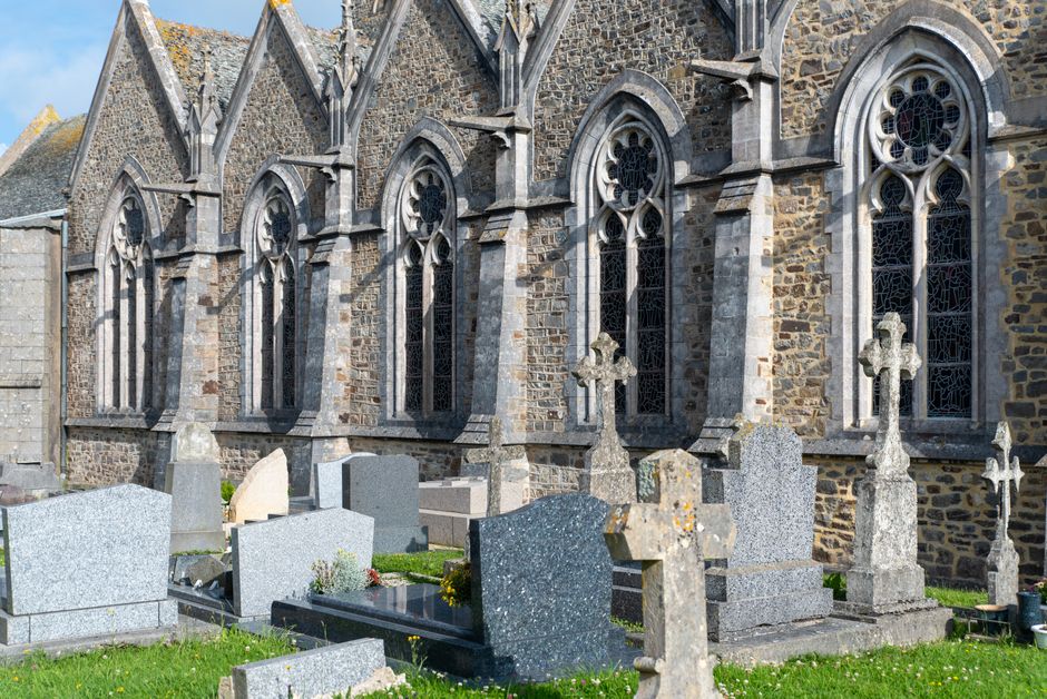 Exterior view of Brompton Cemetery showing the stone walls of a historic church with Gothic-style windows, surrounded by a well-maintained graveyard with various headstones and crosses made of granite and limestone. The area is illuminated by natural daylight, emphasizing the weathered textures of the stone surfaces and the lush green grass in the foreground. Cleaners SW10 specializes in surface cleaning and maintenance, providing deep cleaning and sanitisation services for historic sites and memorials such as those at Brompton Cemetery.