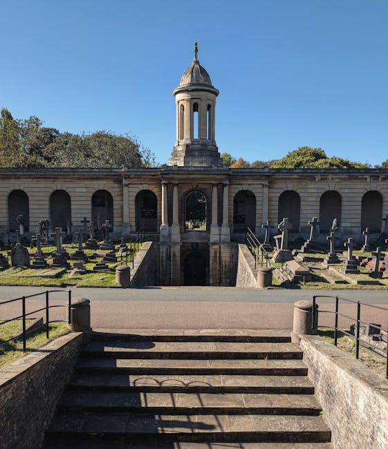 A stone pathway leading up to a memorial structure with a domed cupola, situated within Brompton Cemetery. The pathway is bordered by short metal railings on both sides, and the steps are clean and well-maintained. Surrounding the pathway are rows of gravestones and memorials made of stone and metal, set on a grassy underlay. The scene is illuminated by natural daylight, highlighting the details of the historic site with clear blue sky overhead. The image reflects meticulous maintenance and surface cleaning of the outdoor monument area, consistent with professional care by companies like Cleaners SW10 for ensuring hygiene and preservation of heritage sites.