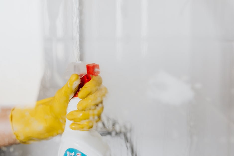 A person wearing bright yellow rubber gloves is holding a white spray bottle with a red nozzle, applying a cleaning solution to a glossy white tiled wall or backsplash. The room appears well-lit with 