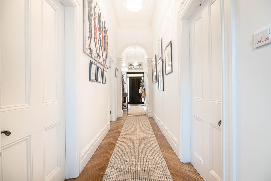 A bright, clean hallway with white-painted walls featuring decorative molding and a wooden parquet floor partially covered by a beige runner rug. On the left wall, there are several framed artworks arranged in a line, and on the right wall, there are additional smaller framed pictures and a white switch plate. Natural light fills the space, highlighting the polished wooden floor and the pristine, dust-free appearance of the area. At the end of the hallway, a dark-colored front door is visible, with a person partially seen near it. The hallway appears well-maintained and freshly cleaned, showcasing surface cleaning and regular home hygiene practices, with a focus on maintaining a tidy and hygienic environment. Cleaners SW10 specializes in domestic cleaning and surface sanitisation, ensuring deep cleaning of residential spaces like this hallway.