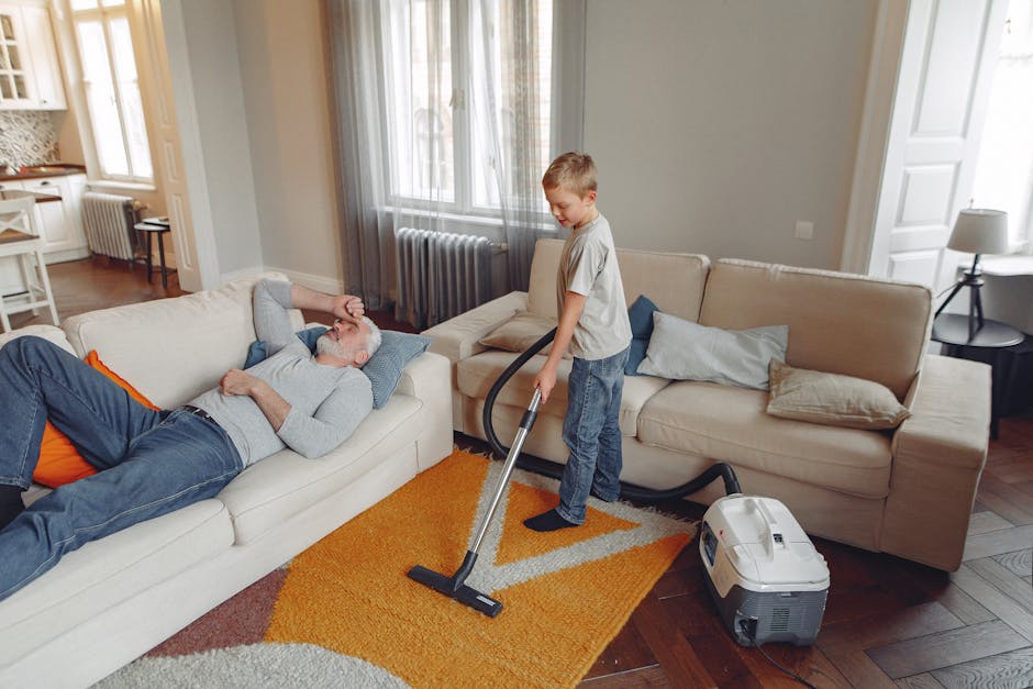 A person kneeling on a patterned carpet while using a yellow and black vacuum cleaner for surface cleaning. The individual is wearing a beige jacket and blue jeans, and is carefully handling the vacuum's hose and filter compartment, which is open to reveal internal components. The room is illuminated with soft lighting, and the carpet appears clean and well-maintained. This scene reflects professional deep cleaning or routine domestic cleaning services provided by Cleaners SW10, specializing in carpet sanitisation and maintenance near Old Brompton Road, SW10.