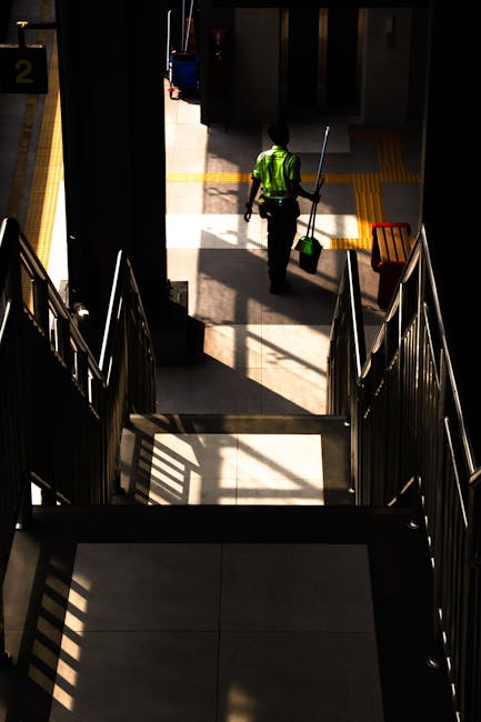 A commercial cleaning worker wearing a high-visibility yellow vest and dark work trousers is seen from the top of stairway, walking across a tiled corridor at West Brompton Station. The worker is holding a green cleaning bucket with a mop handle, and sunlight is casting shadows through large windows onto the floor, highlighting the clean, polished surface. The stairway has metal railings on both sides, and the area appears well-maintained with clear walkways, reflecting professional surface cleaning and sanitisation practices consistent with the services provided by Cleaners SW10 for station and commercial environments.