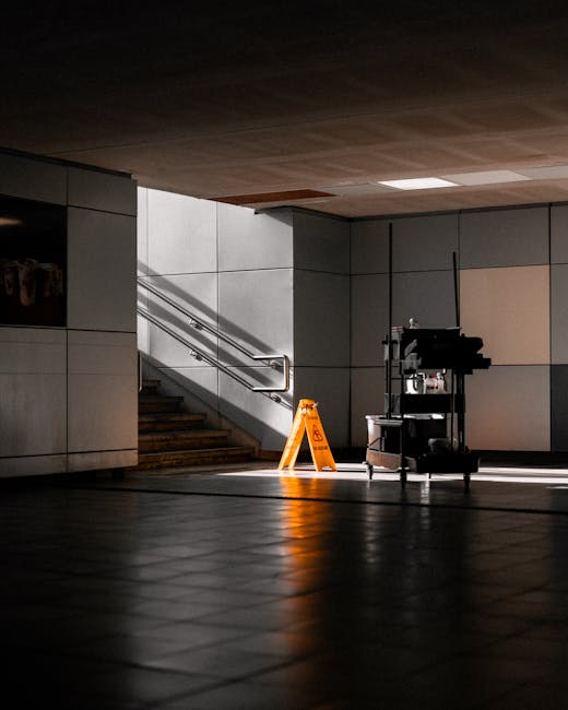 The image depicts a section of an underground station, likely West Brompton Station, with a clean, tiled floor and a staircase leading upward to the platform. The floor surfaces are smooth ceramic tiles that reflect light, indicating a well-maintained, polished condition. To the right of the staircase, there is a modern cleaning trolley equipped with various cleaning supplies, and a tall, black spray machine or vacuum cleaner. An orange safety sign is placed on the floor near the trolley, warning of wet surfaces, which suggests ongoing or recent cleaning activity. The station's wall surfaces are composed of large, light-colored panels with minimal visible grime, and the area is illuminated by artificial lighting, creating a balanced contrast with the darker, shadowed floor. This scene highlights professional deep cleaning and sanitisation efforts typical for public transport stations, with Cleaners SW10 providing thorough surface cleaning and hygiene maintenance in such environments, aligning with their mention on the West Brompton Station cleaning guide for landlords, SW10.